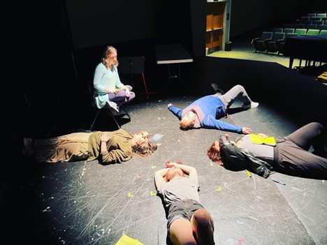 Photo of students lying on the floor in a circle with one student kneeling near one of the students. London Thorn, Suzy Bounsavy, Edward Mountz, Blue Chronister, and Nikki Phillips are the students rehearsing for an upcoming play.